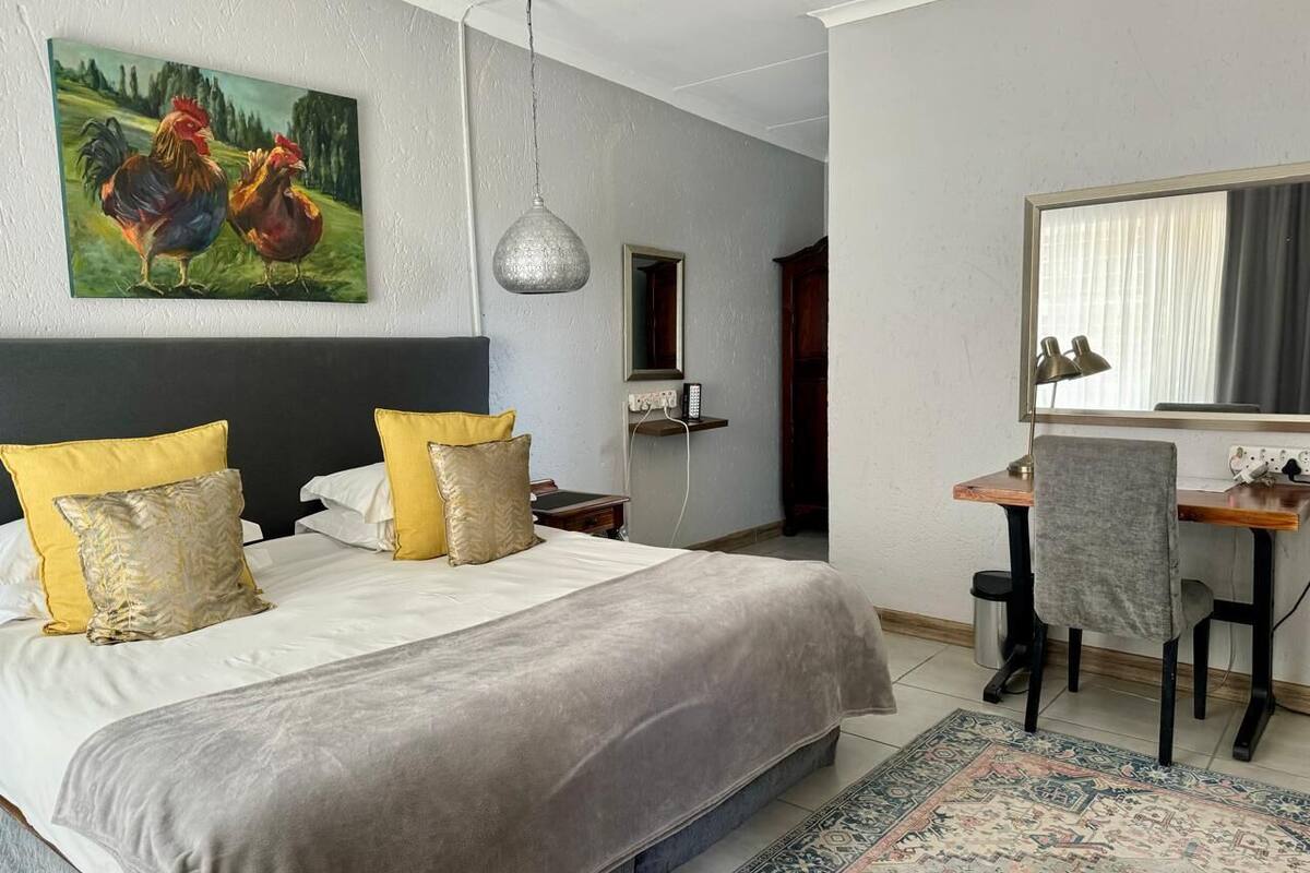 A bedroom with a large bed, two bedside tables, a desk with a chair, a hanging lamp, and a painting of roosters.
