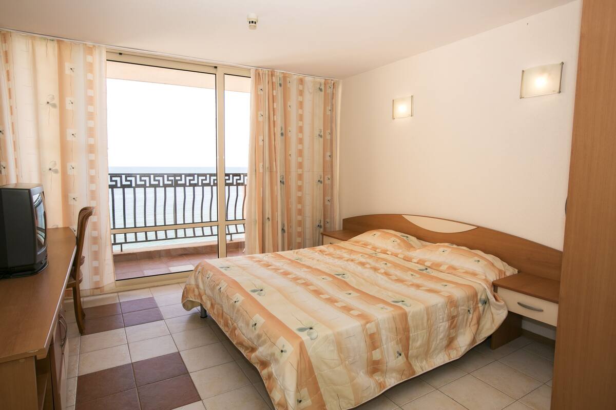 A hotel room with a bed, a desk, a chair, a TV, and a balcony with a railing.