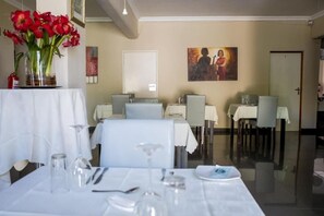 A dining area with tables set for two, a vase of red flowers, and a painting on the wall. - Ruby Stone Boutique Hotel (Polokwane)
