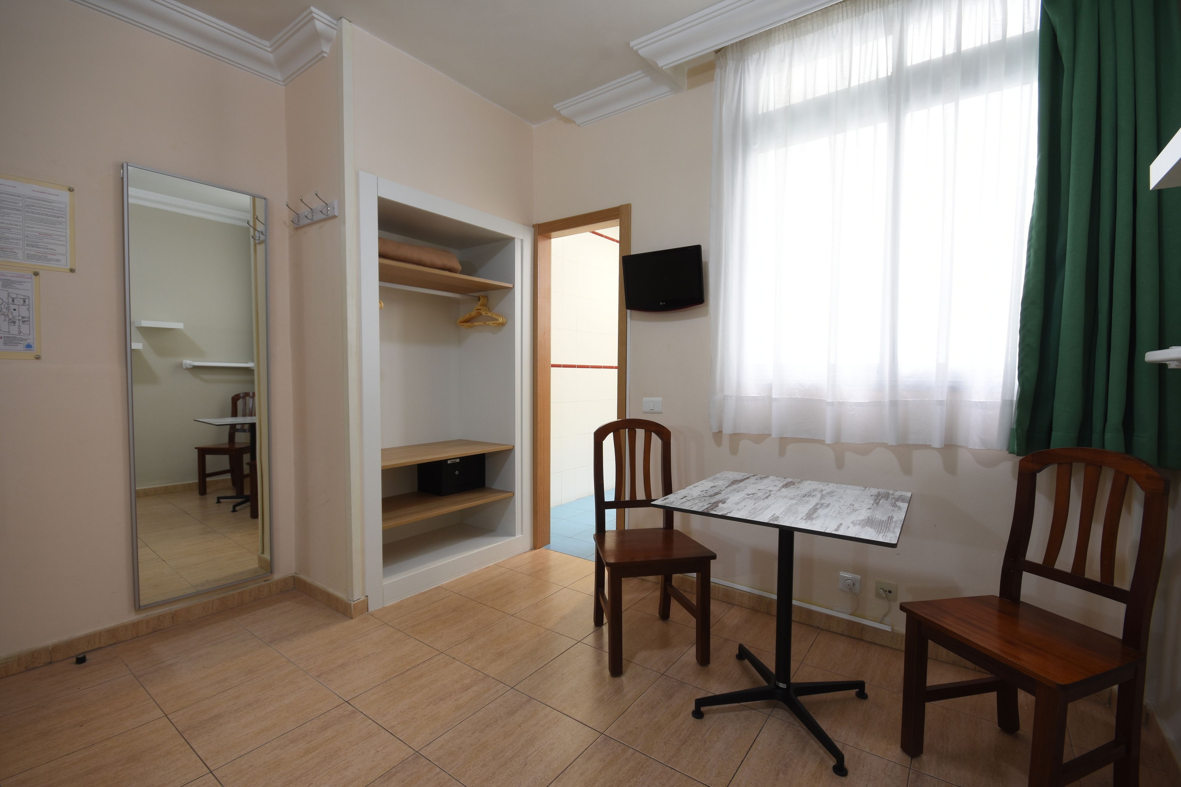Una habitación con piso de baldosas, una silla de madera, una mesita, un espejo, un armario y una ventana con cortinas.