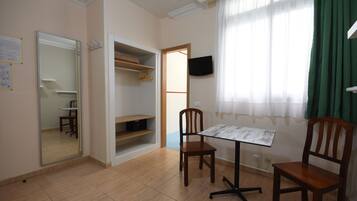 Una habitación con piso de baldosas, una silla de madera, una mesita, un espejo, un armario y una ventana con cortinas.