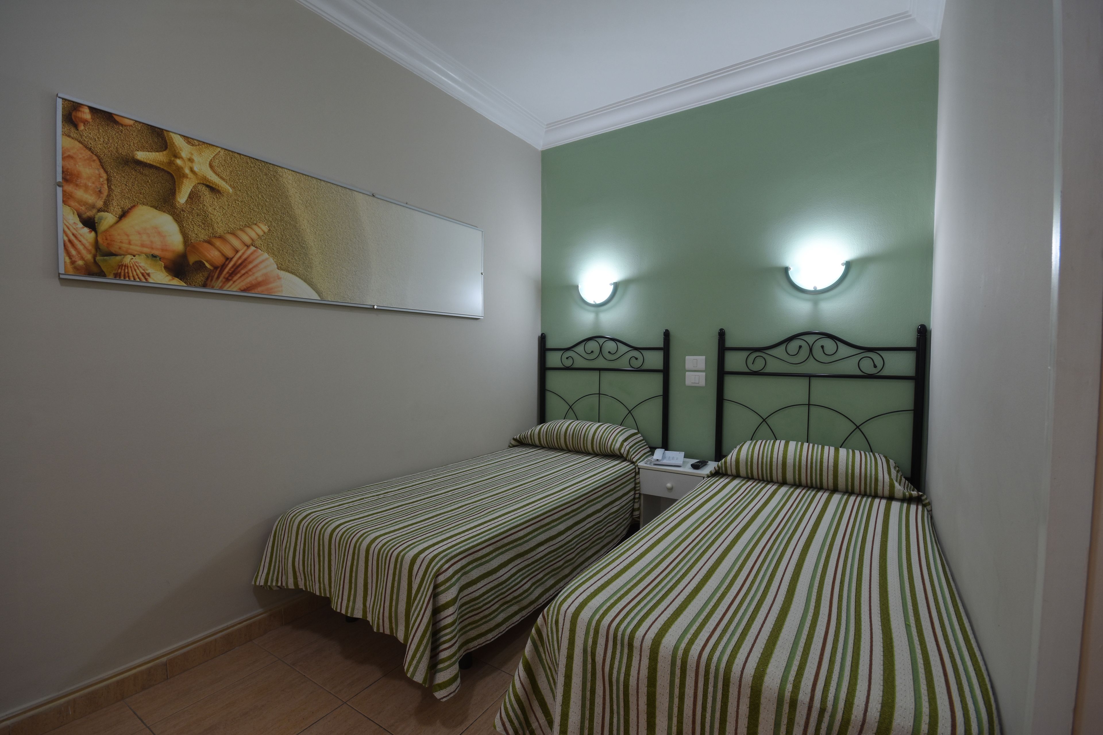 Quarto com duas camas de solteiro, roupa de cama listrada, luminária fixada na parede e um quadro com conchas.