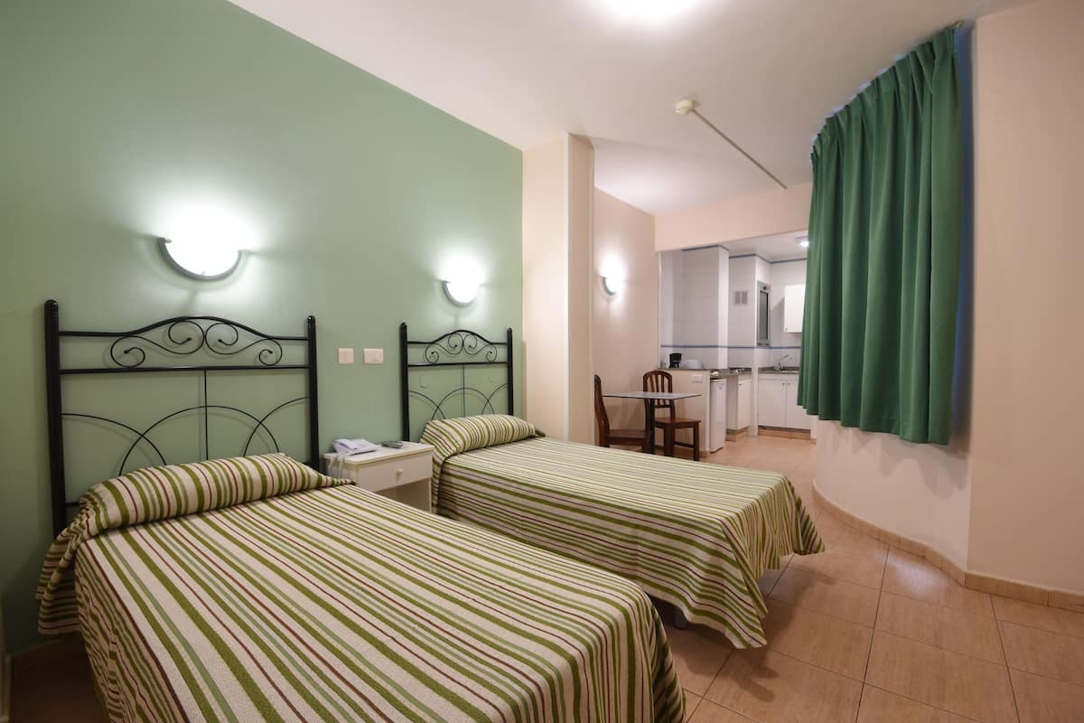 A room with two single beds, a small table, and a chair. There is a kitchenette in the background.