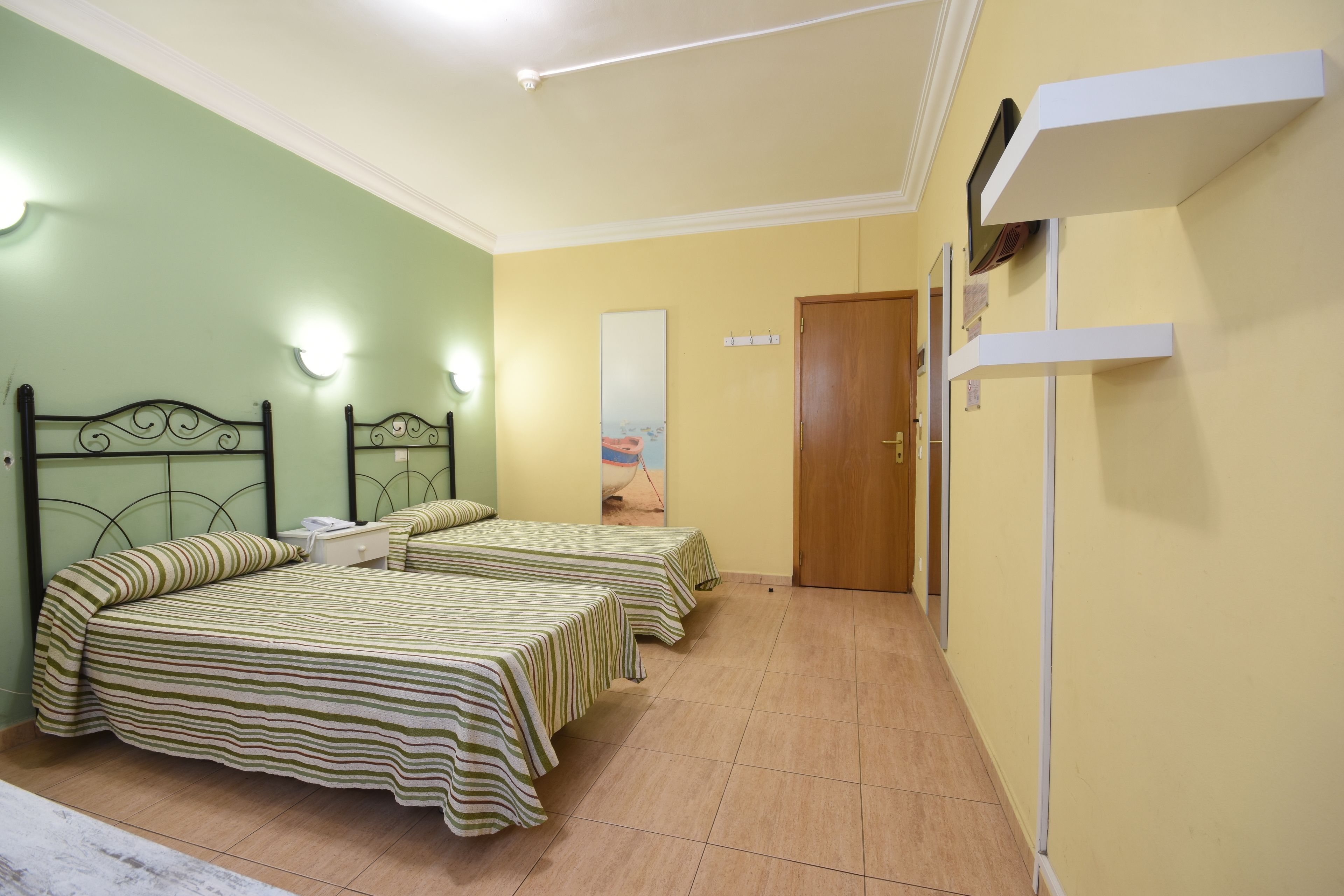 Una habitación con dos camas, una puerta de madera y un estante blanco con un espejo.
