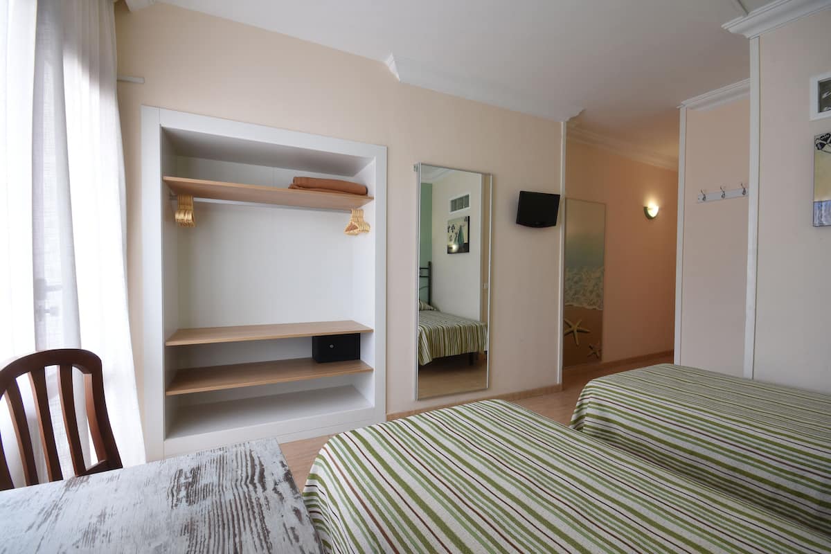 A bedroom with a bed, a chair, a table, a wardrobe, a mirror, and a wall-mounted TV.