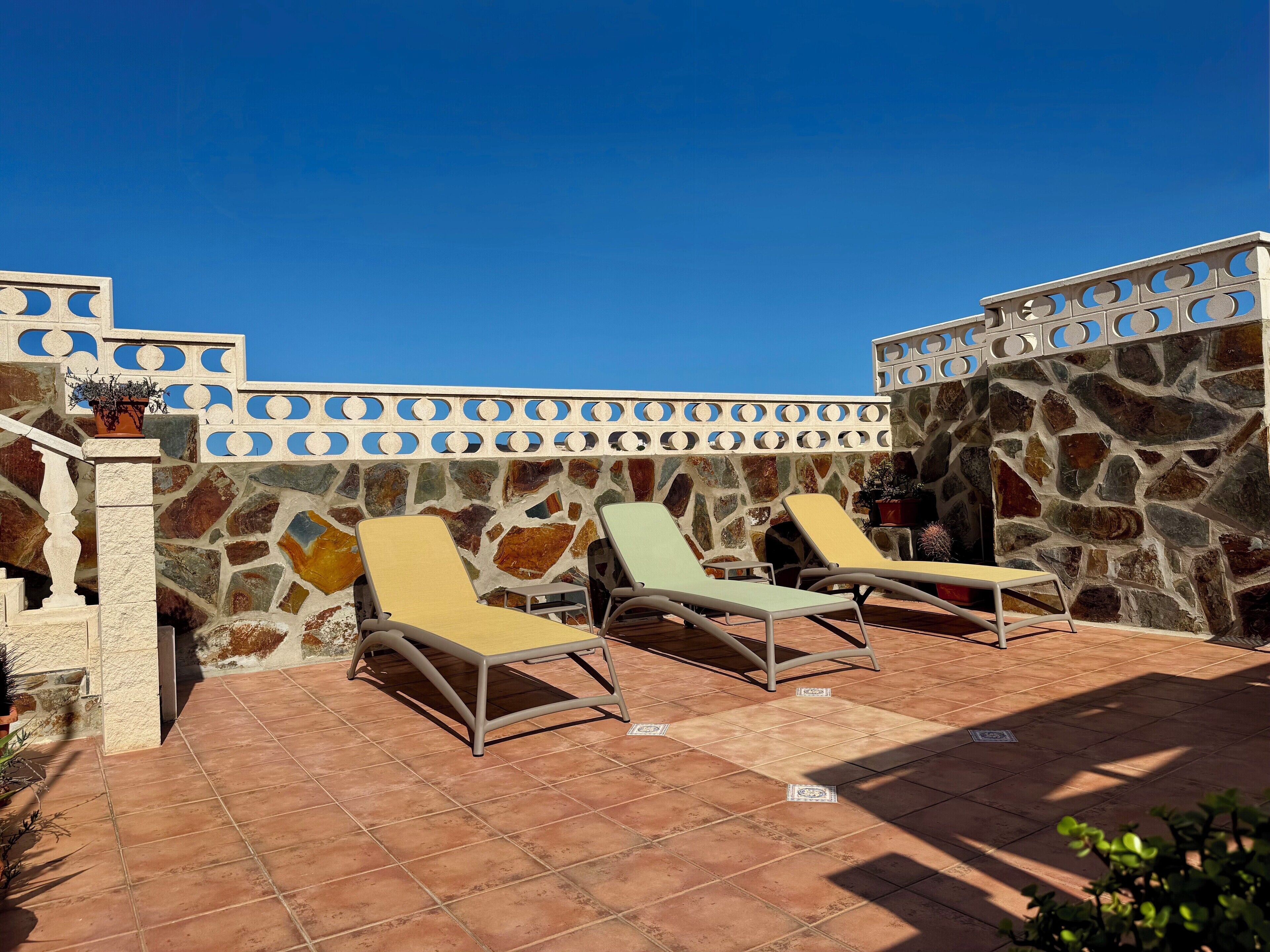 A rooftop terrace with stone walls, two lounge chairs, and a potted plant.
