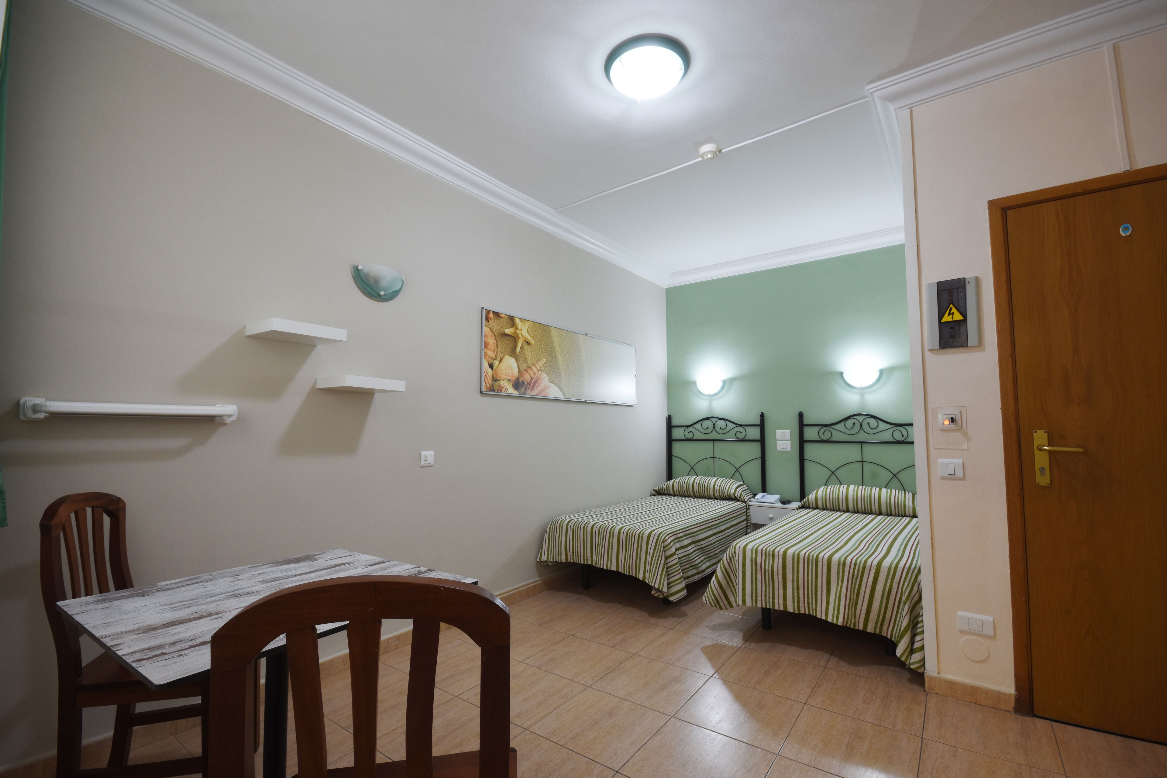 Un dormitorio con dos camas individuales, una silla de madera, una mesa, un estante en la pared y una puerta.