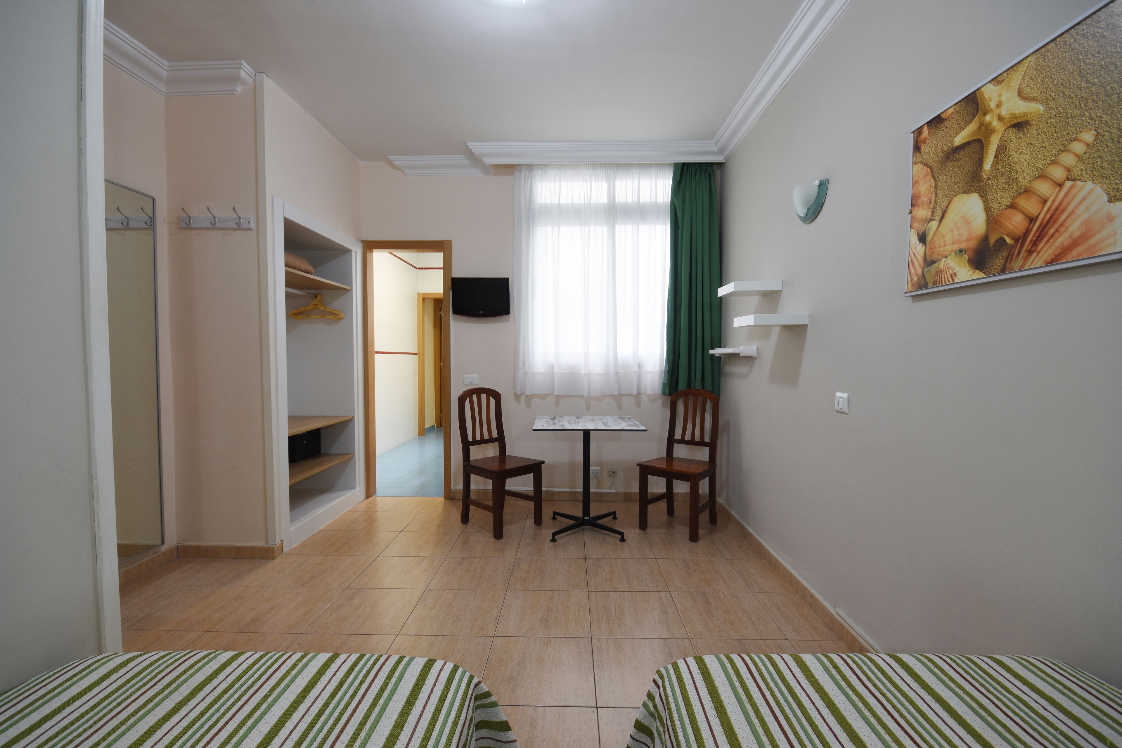 Una habitación con dos camas, una mesita, dos sillas, un televisor y un armario.