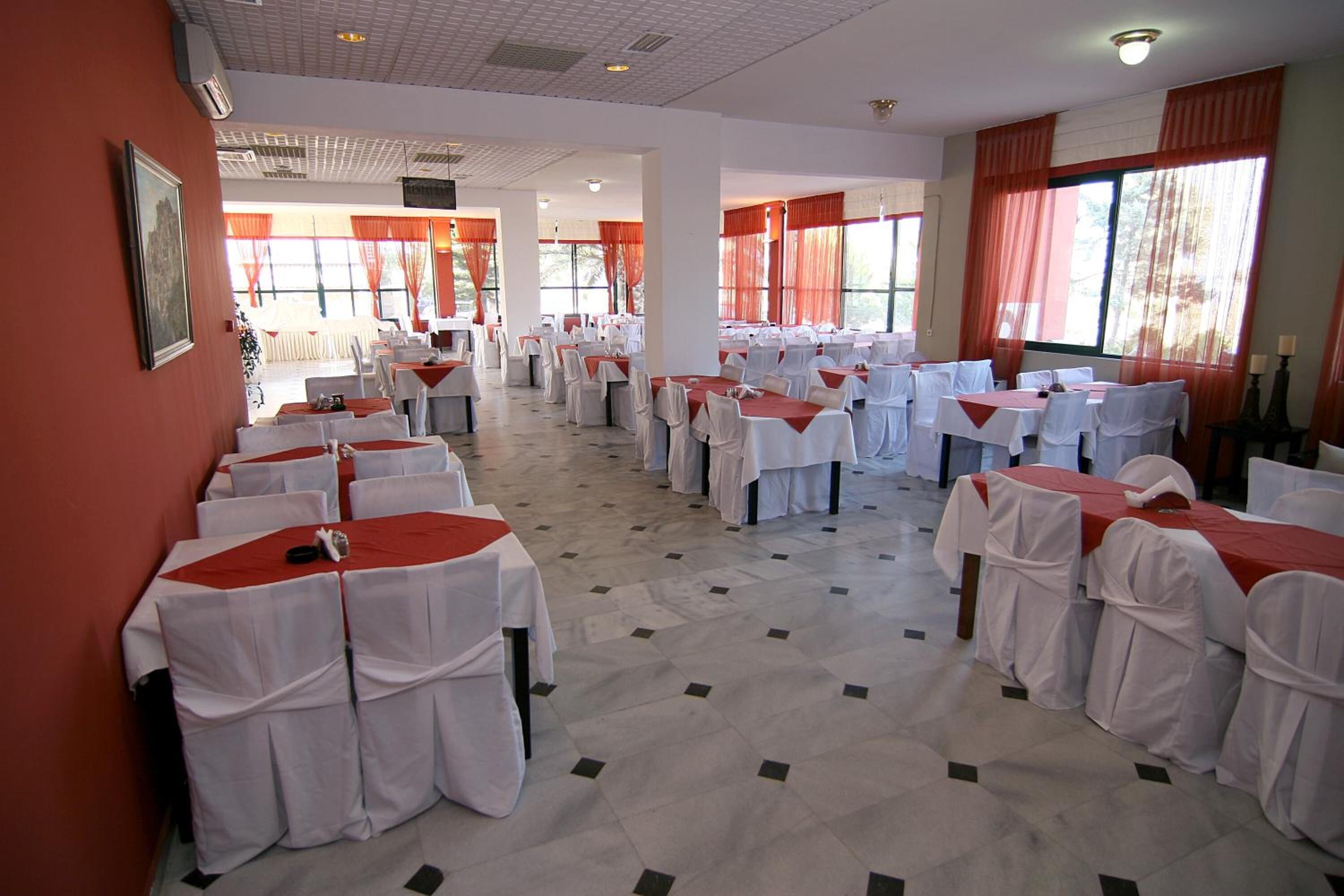 Un salón de banquetes con filas de mesas cubiertas con manteles blancos y caminos de mesa rojos, ventanas amplias con cortinas rojas y piso de baldosas.