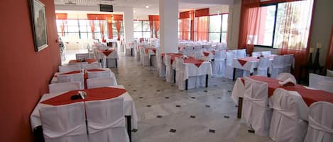 Un salón de banquetes con filas de mesas cubiertas con manteles blancos y caminos de mesa rojos, ventanas amplias con cortinas rojas y piso de baldosas.