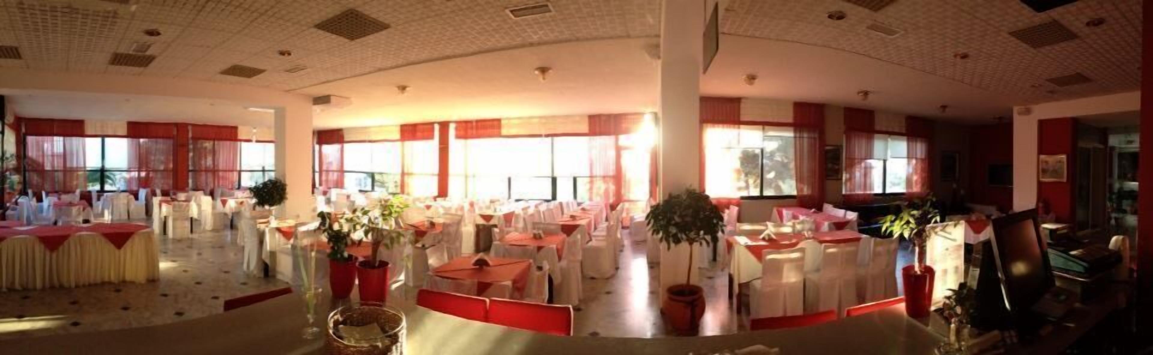 Un vestíbulo amplio con varias mesas redondas y sillas, ventanas grandes con cortinas rojas y un techo alto con patrones tipo cuadrícula.