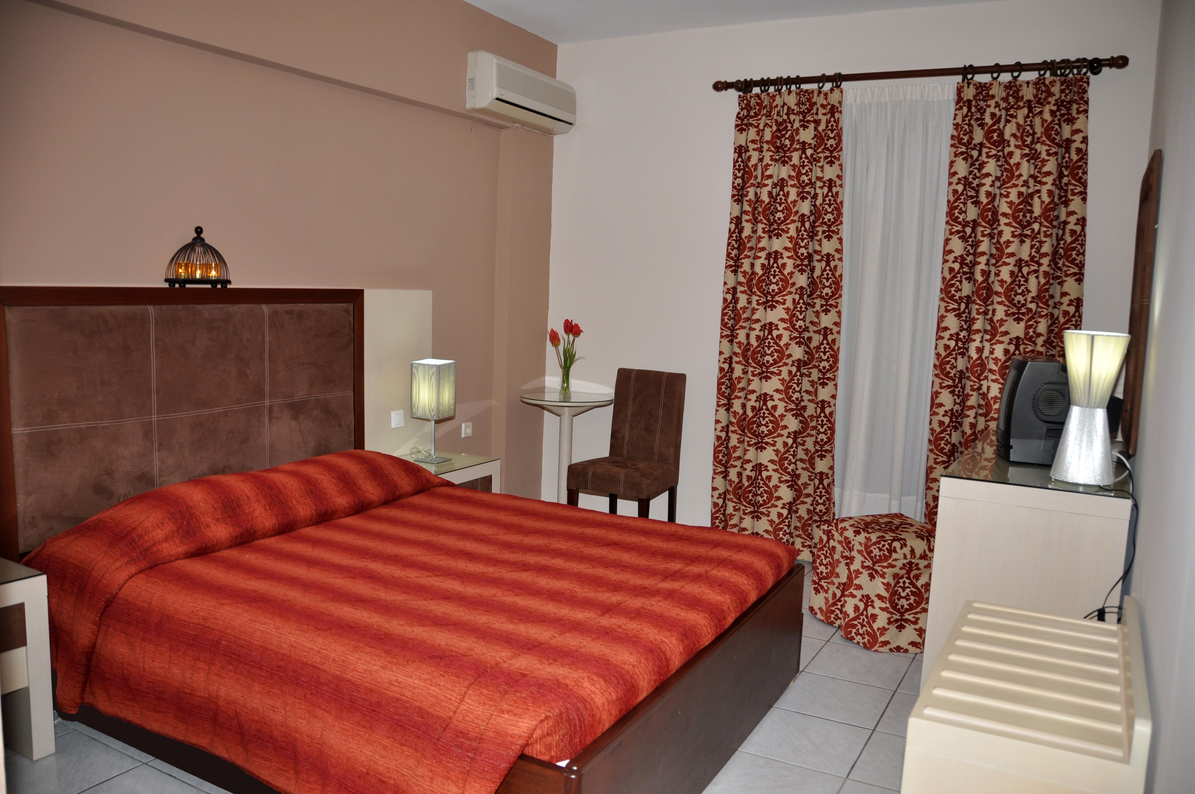 Habitación de hotel con una cama grande, un escritorio con lámpara, una silla, una mesita con un jarrón de flores y un radiador.