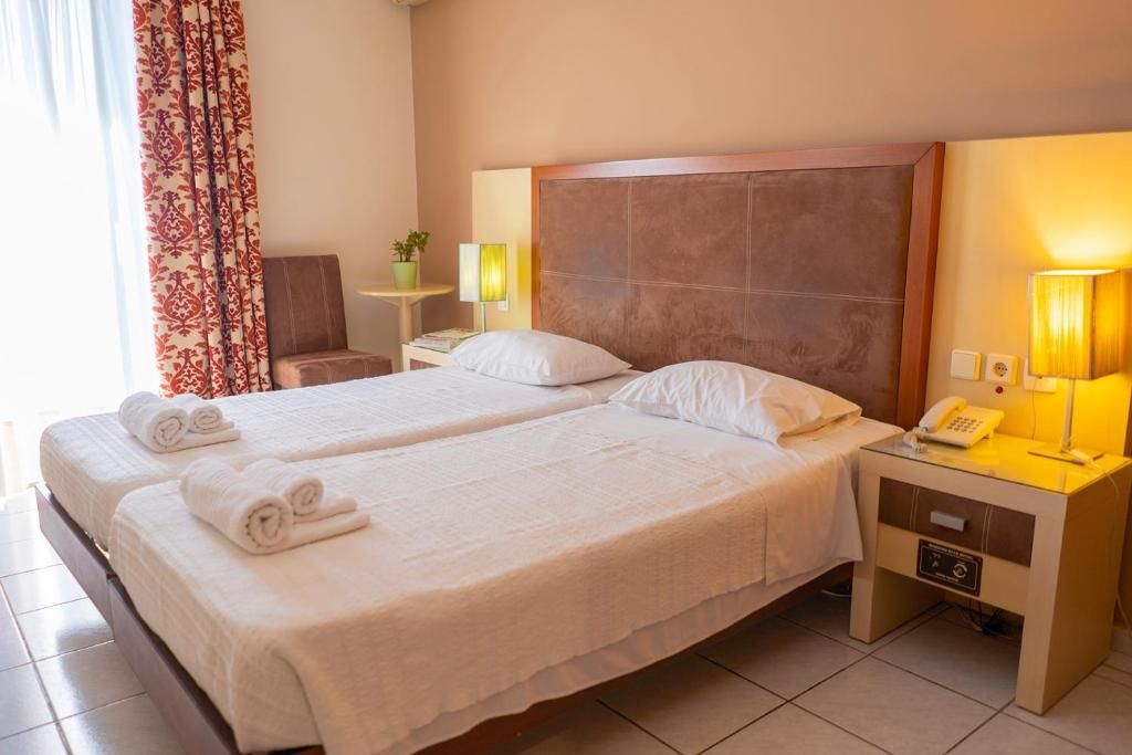 Habitación de hotel con dos camas, una mesita de noche con lámpara, una silla y un ventanal con cortinas.