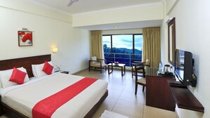 A hotel room with a large bed, a desk with a TV, a chair, and a view of the mountains through the window. - Clouds Valley Leisure Hotel (Devikolam)