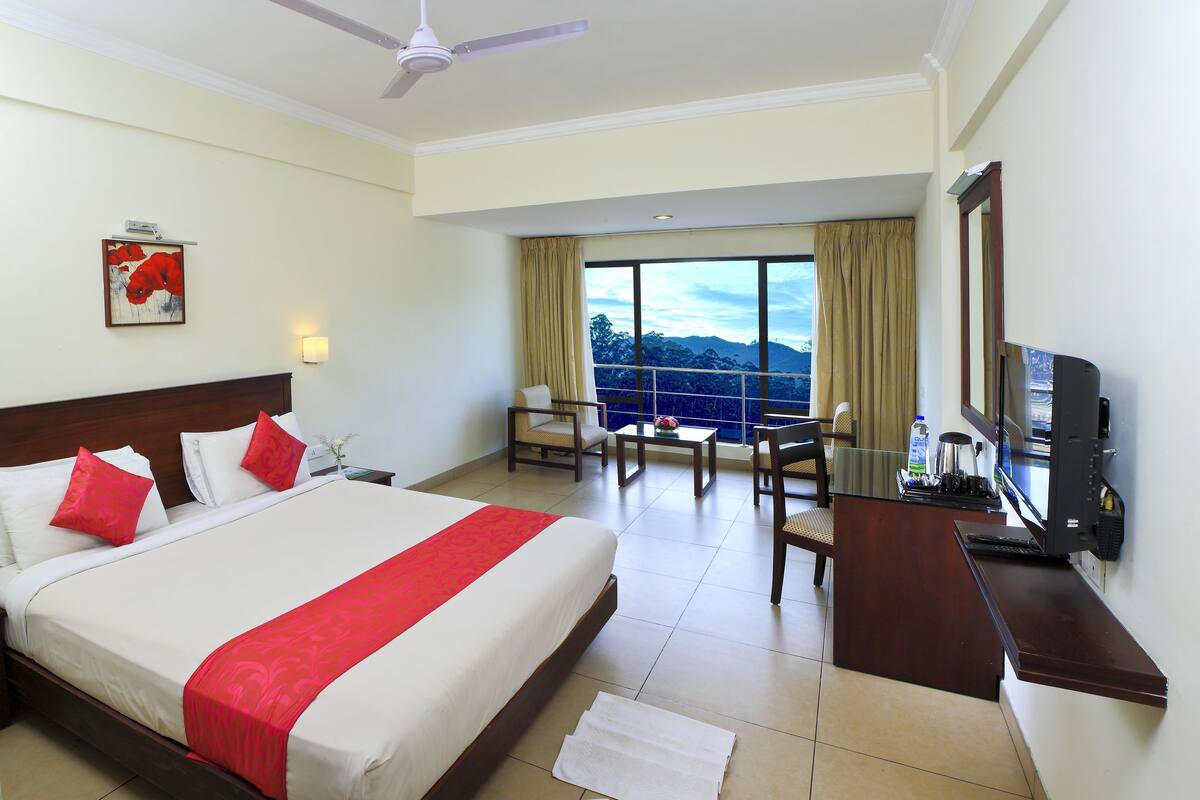 A hotel room with a large bed, a desk with a TV, a chair, and a view of the mountains through the window.
