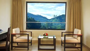 A room with two chairs, a small table with a vase, and a large window with a view of green hills. - Clouds Valley Leisure Hotel (Devikolam)
