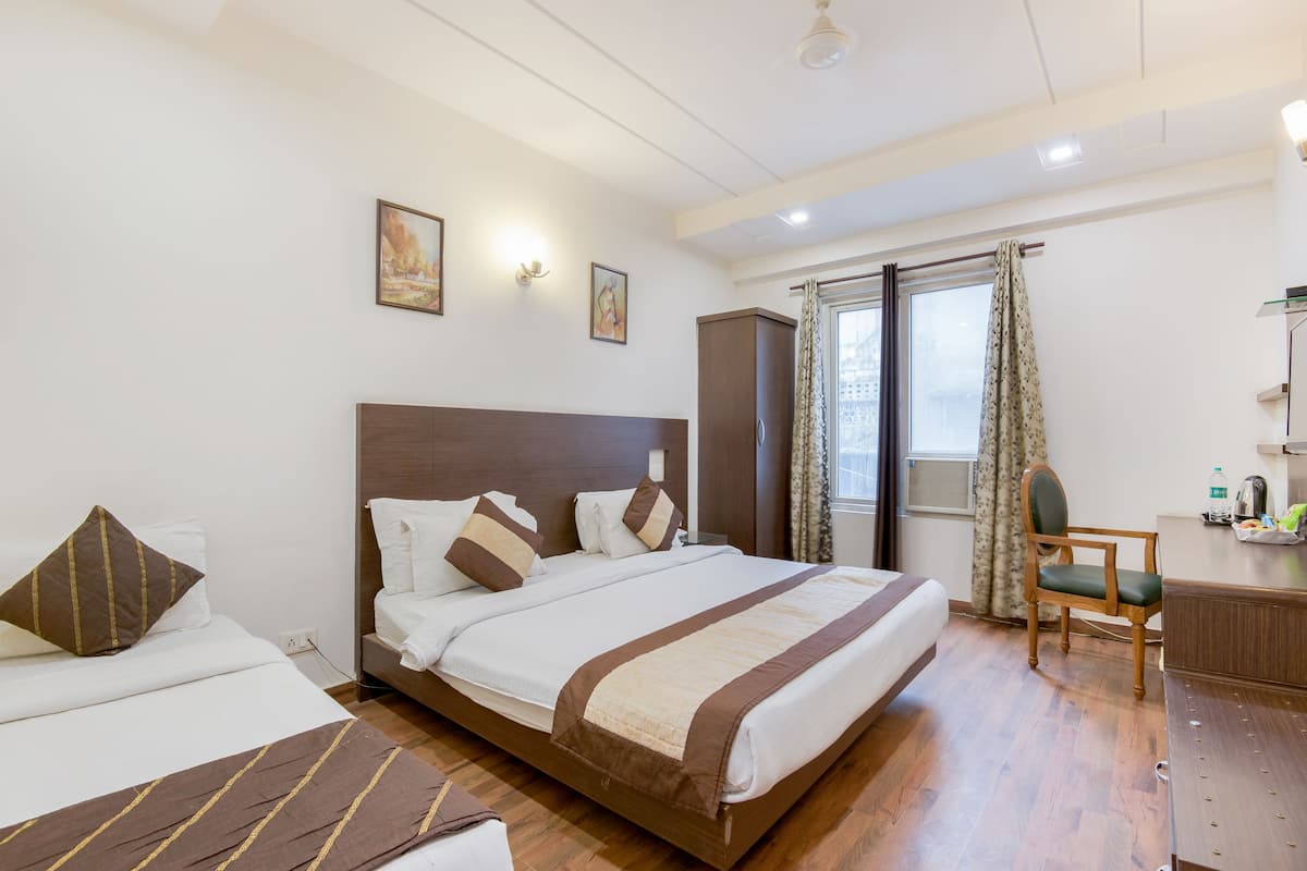 A hotel room with a large bed, a desk, and a chair. There are two paintings on the wall and a window with curtains.
