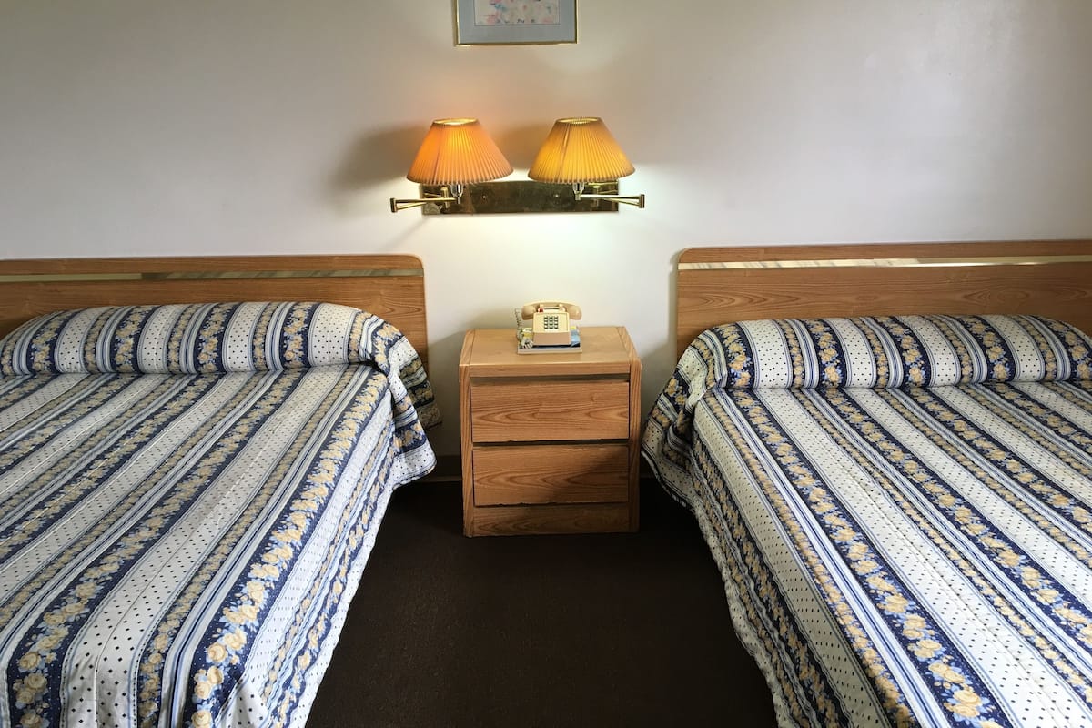 A room with two beds, each with striped bedding, a wooden nightstand with a telephone, and two wall-mounted lamps.