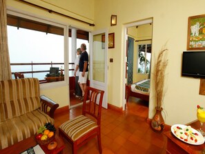 A hotel room with a balcony, a bed, a TV, and a dining table with fruit. - Blackberry Hills Munnar - Nature Resort & Spa (Devikolam)