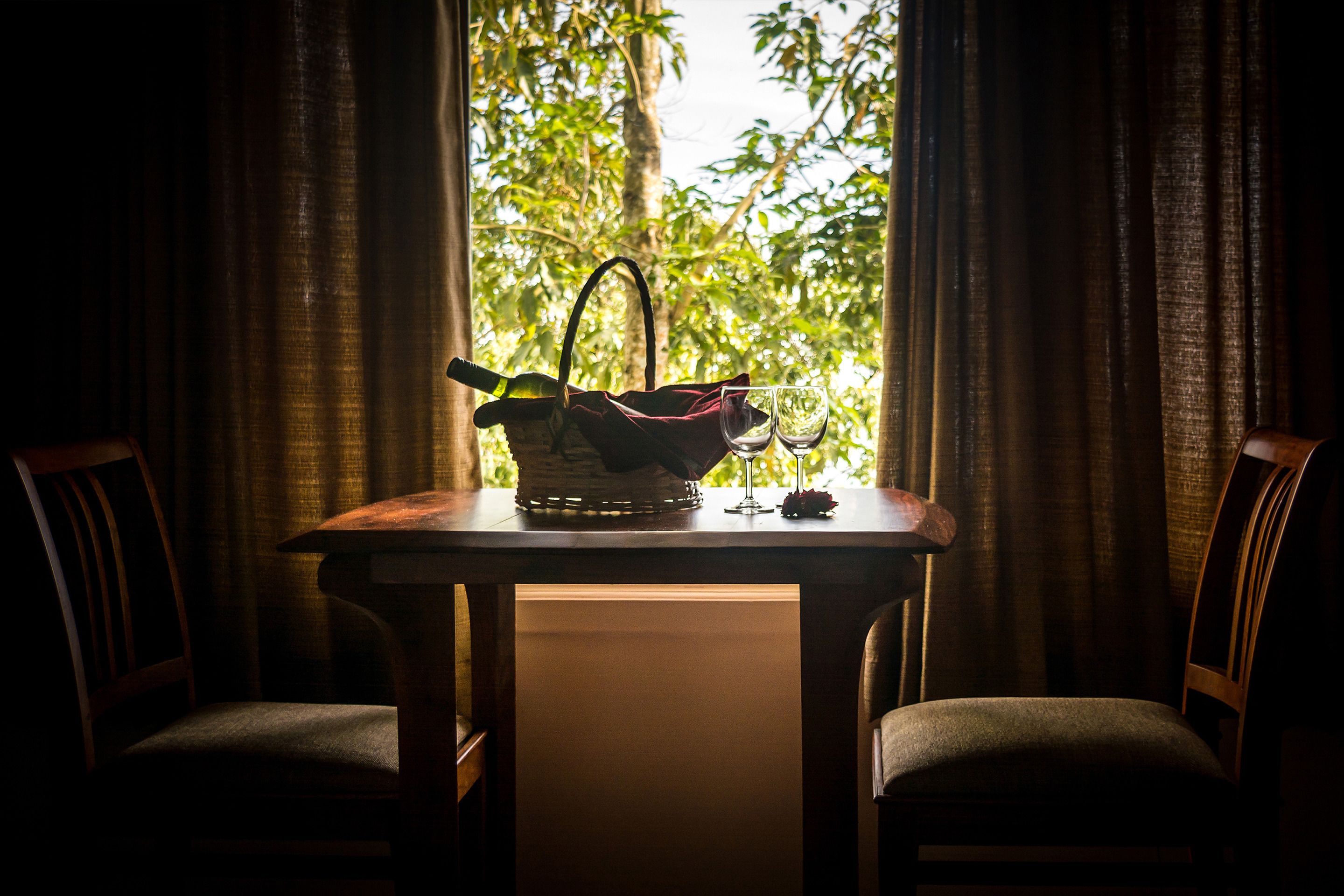 A table with a wicker basket, a wine glass, and a bottle, set against a window with curtains.