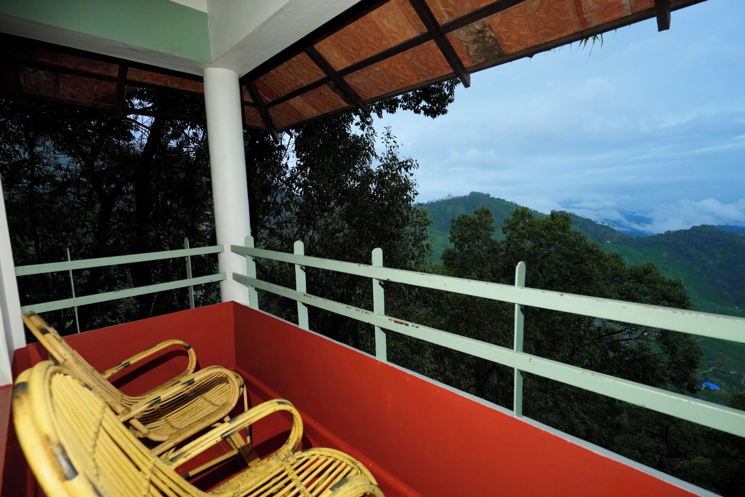 A balcony with wicker chairs, a red wall, and a view of a green landscape with trees and hills.