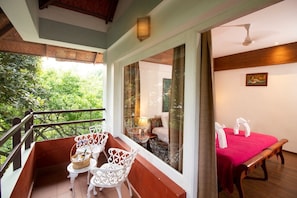 A balcony with white chairs and a table, a bed with a red blanket, and a view of greenery. - Blackberry Hills Munnar - Nature Resort & Spa (Devikolam)
