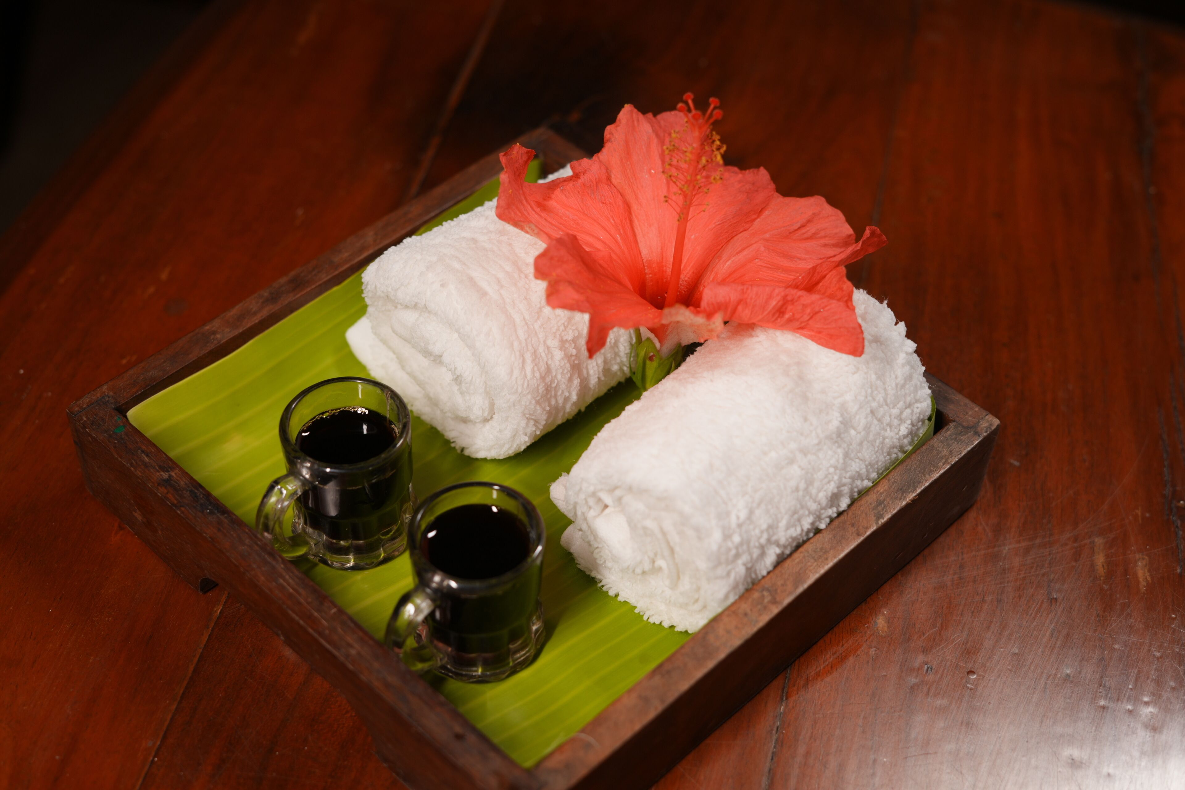 A tray with two white towels, a red hibiscus flower, and two small glasses of tea on a green leaf.
