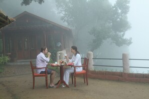 Two people sitting at a wooden table outdoors with a rustic building in the background. - Blackberry Hills Munnar - Nature Resort & Spa (Devikolam)