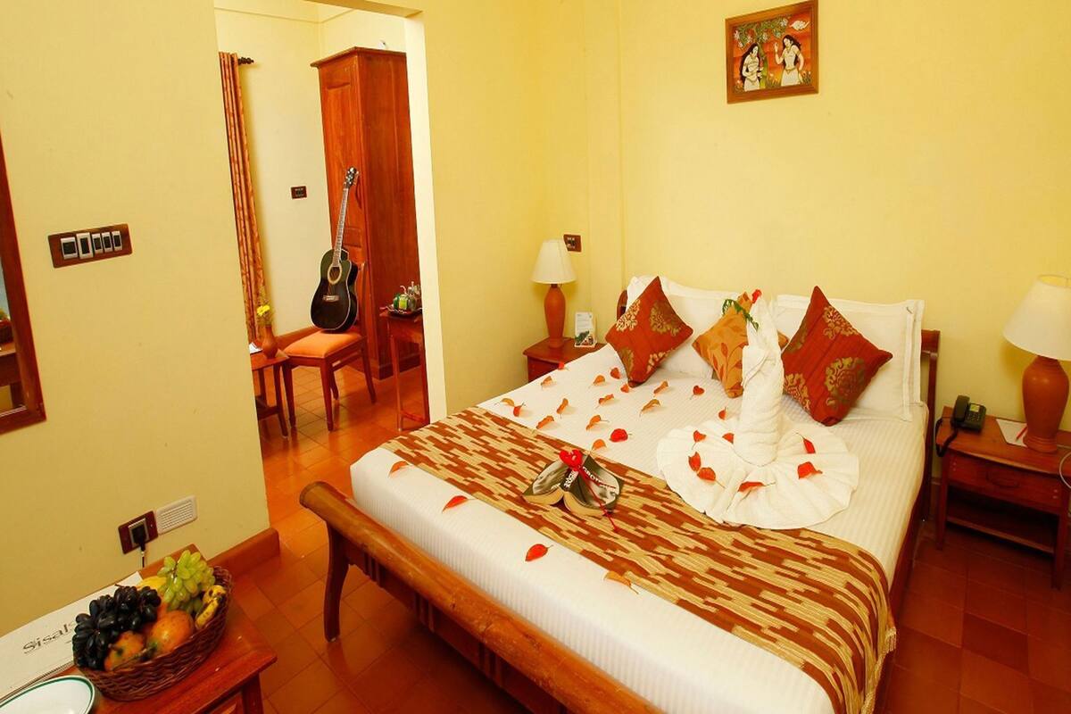 A hotel room with a bed, a desk with a fruit basket, a guitar, and a painting on the wall.