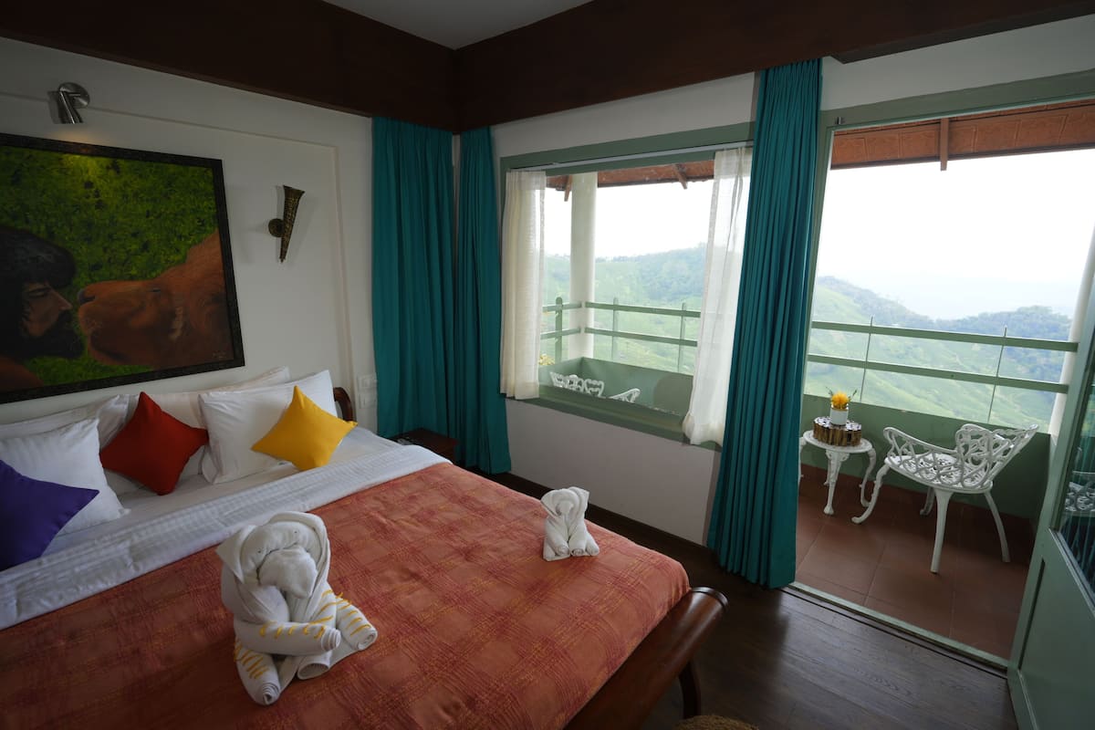 A hotel room with a bed, pillows, a painting, a balcony with a table and chair, and a view of the mountains.