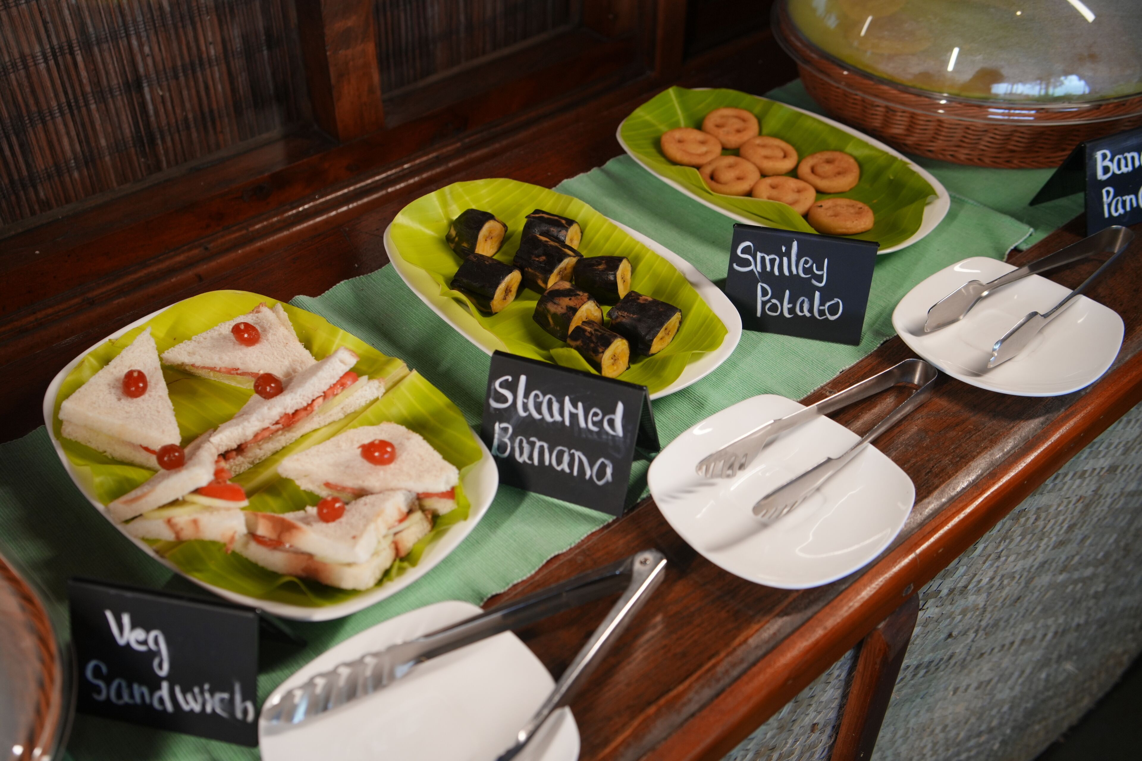 Un bufé con platos identificados, incluyendo sándwich de vegetales, plátano al vapor y papa sonriente.