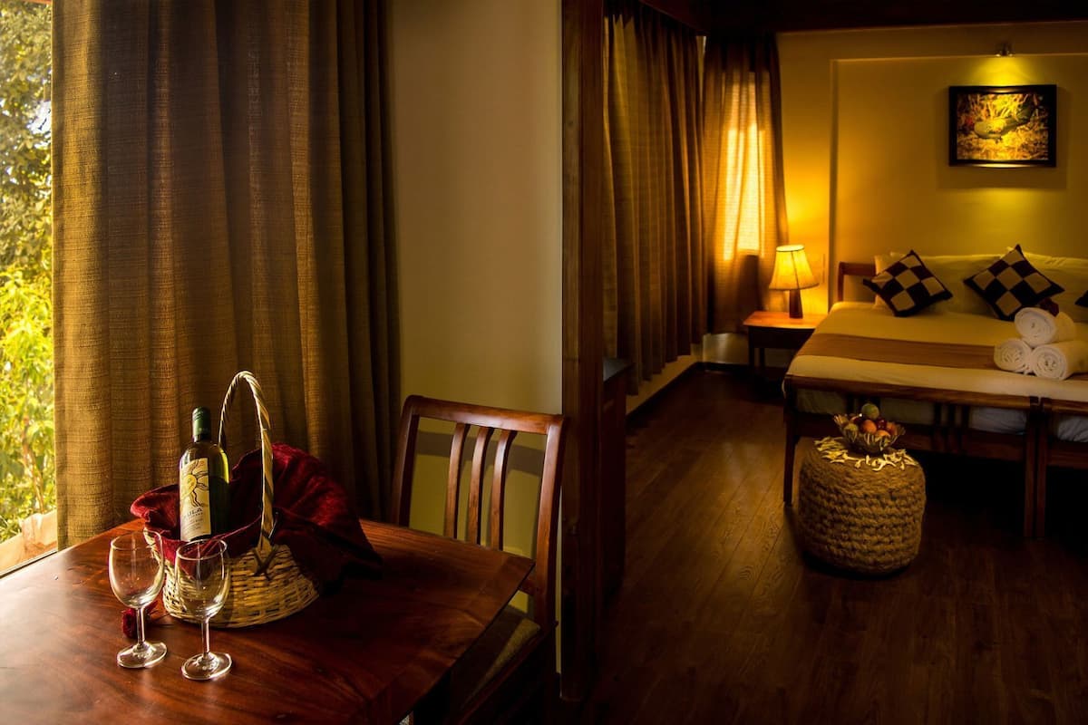 A hotel room with a wooden table, two wine glasses, a bottle of wine, a basket, a bed with checkered pillows, a lamp, and a painting on the wall.