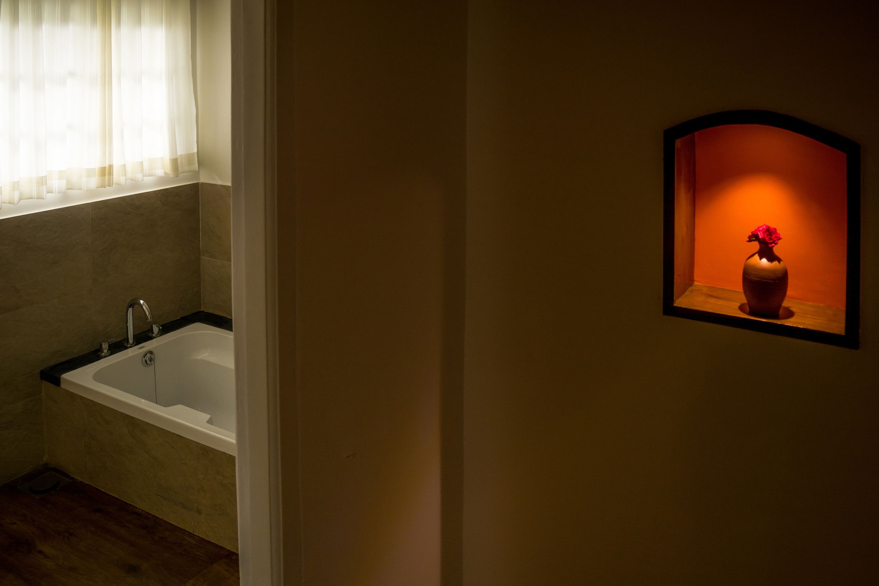 Un baño con bañera, una ventana con cortinas traslúcidas y un florero con flores fijado a la pared.