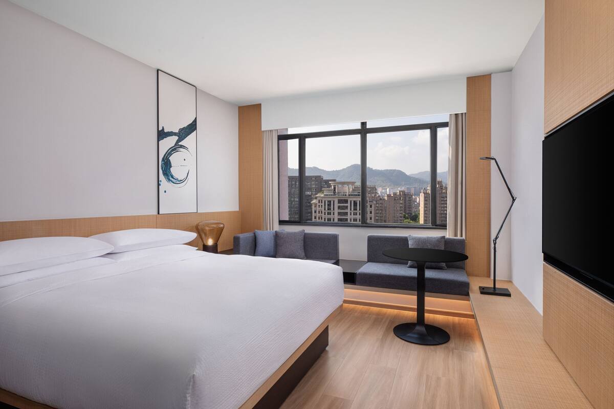 Una habitación de hotel moderna con una cama grande, una zona de estar con una mesa, un televisor y vistas a la ciudad a través de la ventana.