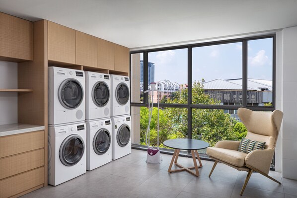 Een moderne wasruimte met gestapelde wasmachines en drogers, een klein tafeltje en een comfortabele stoel, met uitzicht op groen en gebouwen door grote ramen.