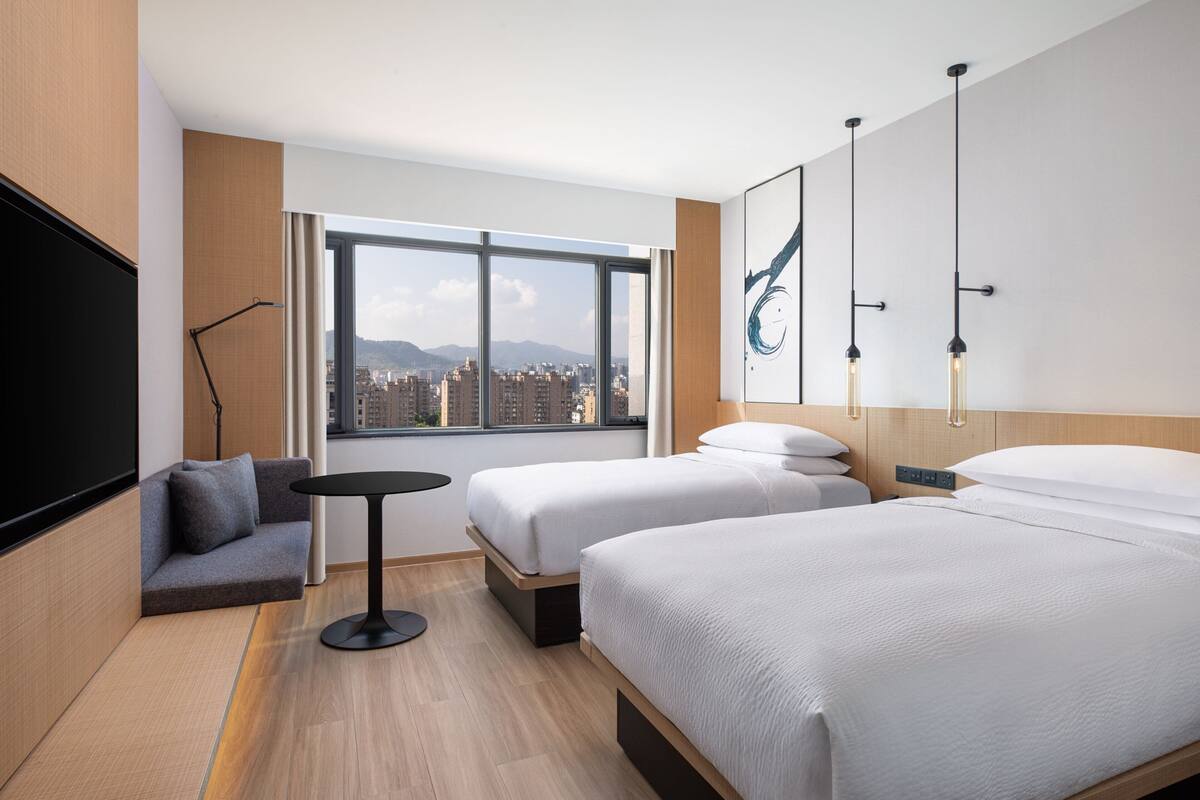 Habitación de hotel con dos camas, un sofá, una mesita, un televisor y un ventanal con vistas a la ciudad.