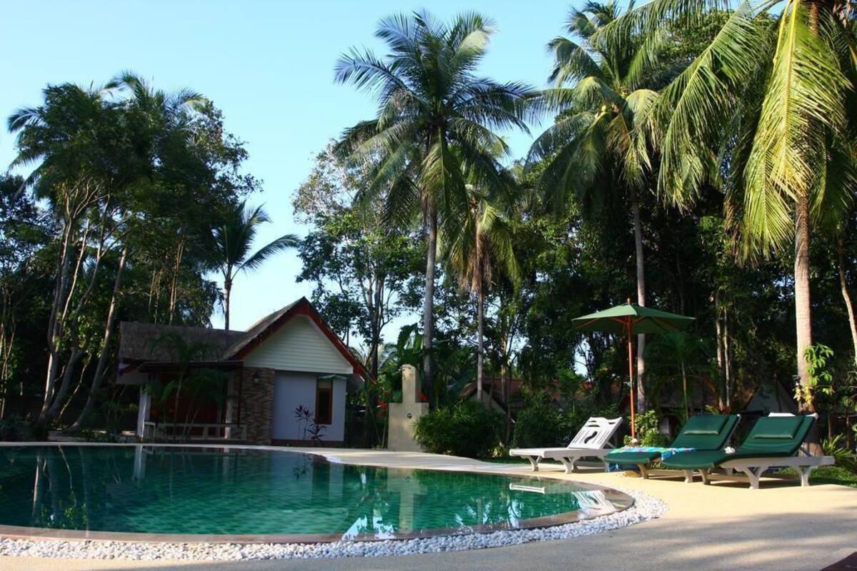 Una piscina rodeada de palmeras, un edificio pequeño y sillas de descanso.