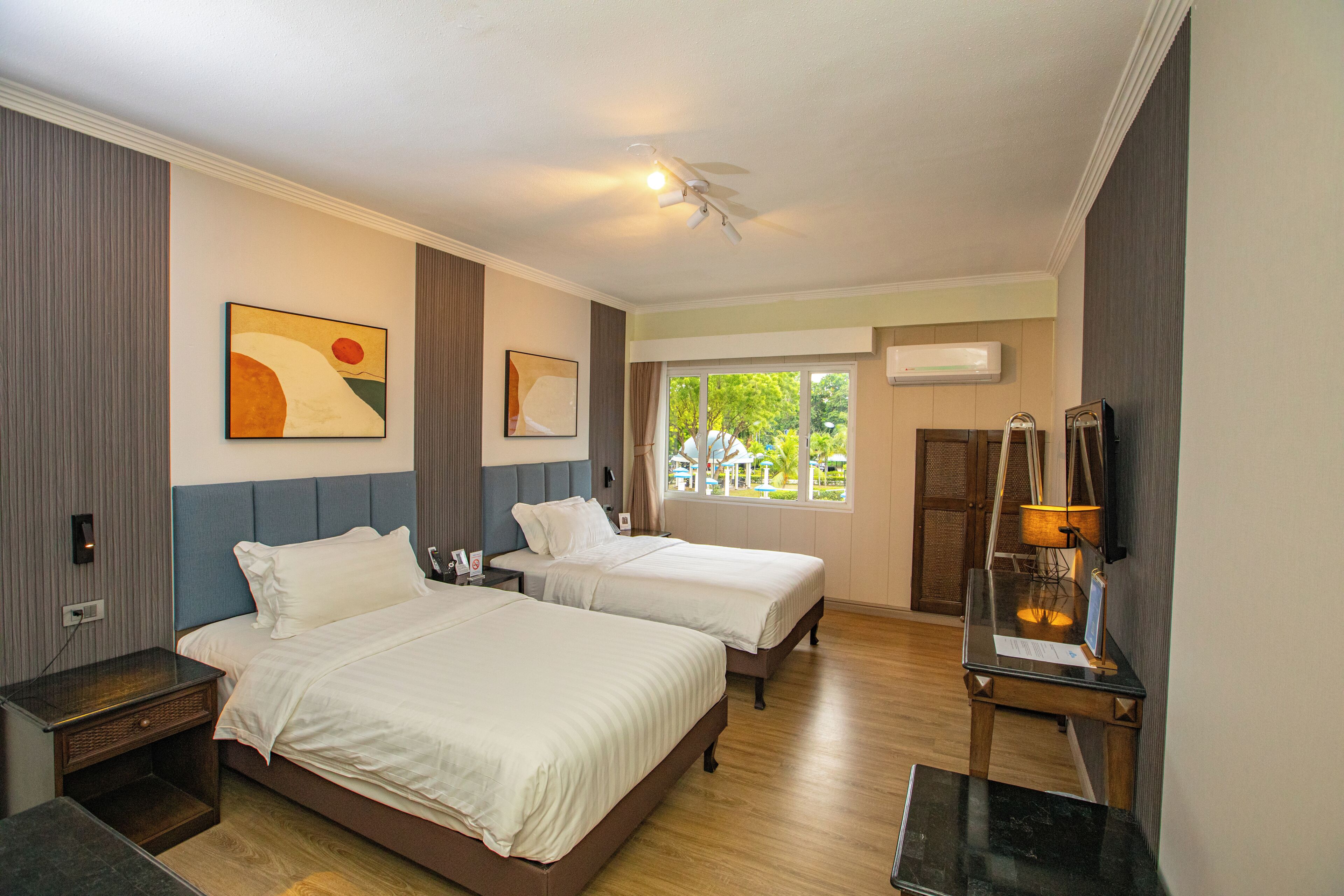 Habitación de hotel con dos camas, un escritorio, ventilador de techo, obras de arte en las paredes y ventana con vistas a zonas verdes.