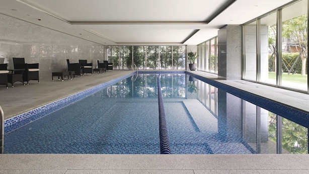 A modern indoor swimming pool with clear glass walls, a tiled floor, and black lounge chairs. - Gloria Residence (Taipei)