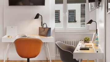 Ein moderner Arbeitsraum mit Schreibtisch, zwei Stühlen, einem Laptop, einer Handtasche, einer Lampe, einem Heizkörper und einem Fenster mit Blick auf Gebäude.