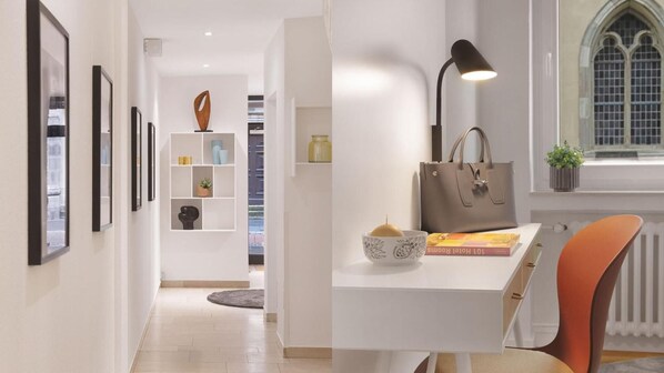A modern hotel room with a white desk, a chair, a lamp, a window with a view, and a bookshelf with decorative items. - Beethoven Hotel Dreesen - furnished by BoConcept (Bonn)