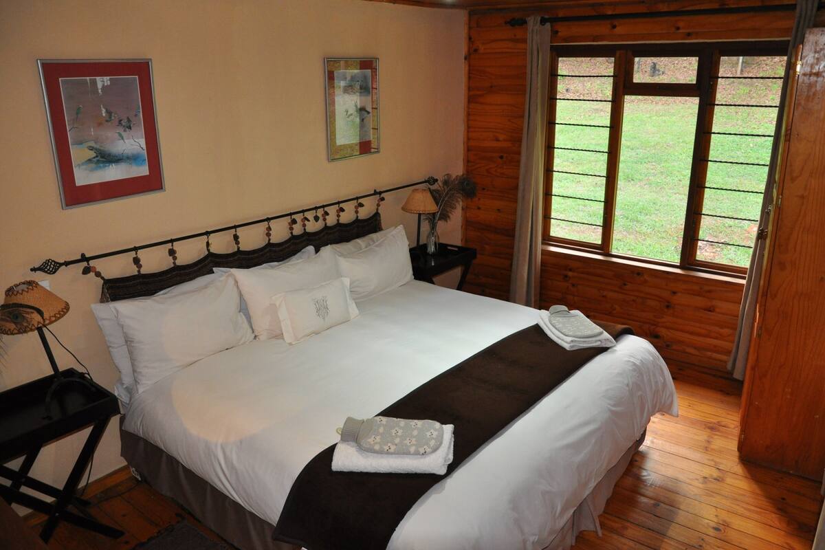 A neatly made bed with white linens, a dark headboard, and a window with a view of a grassy area.