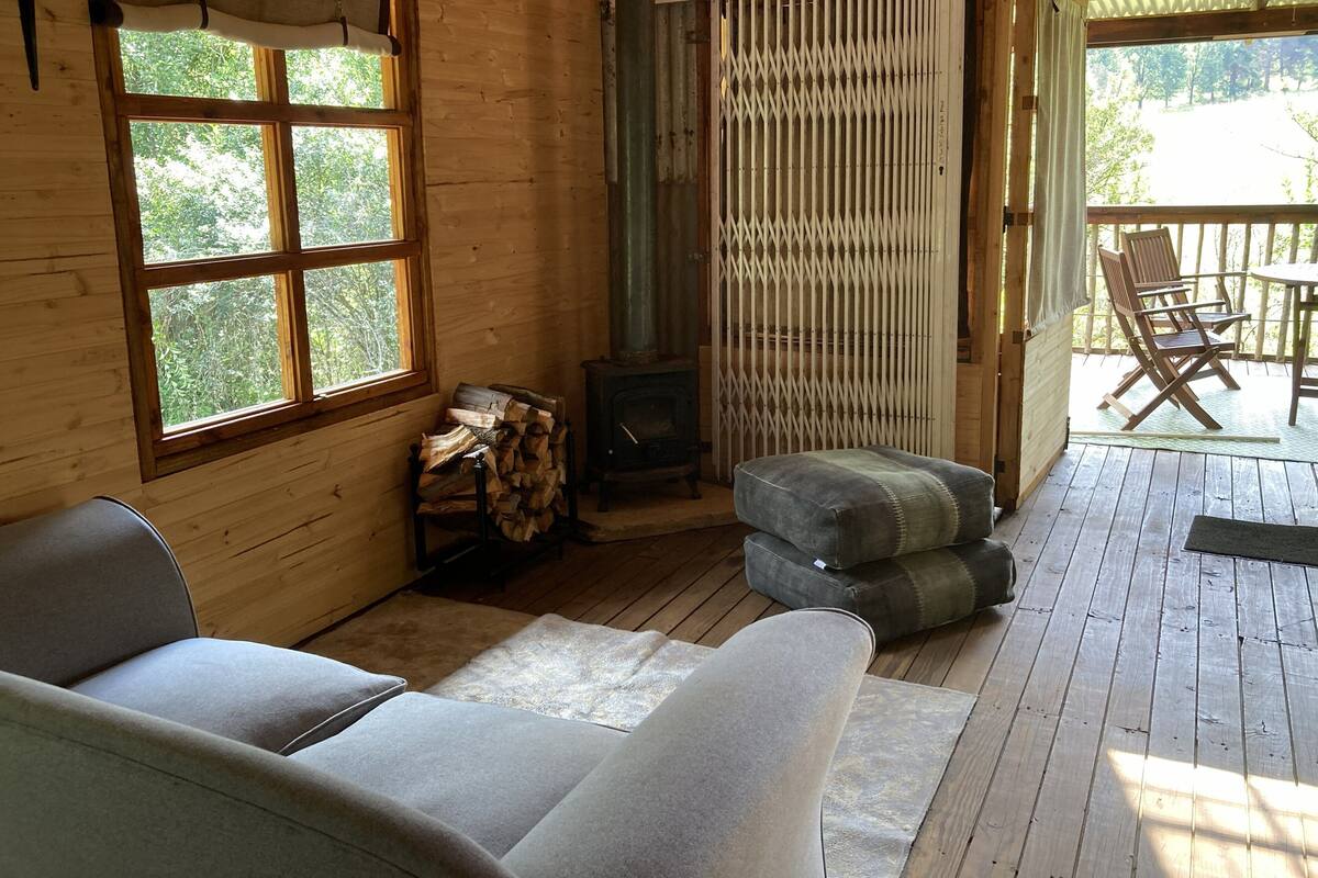 A wooden cabin interior with a sofa, a fireplace, a window with curtains, and a deck area with chairs.