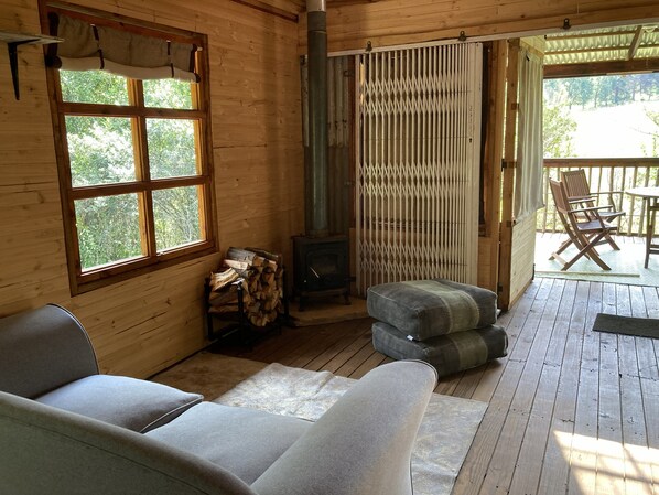 A wooden cabin interior with a sofa, a fireplace, a window with curtains, and a deck area with chairs.