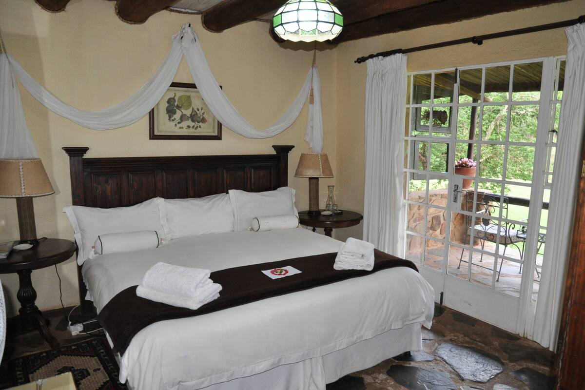 A bed with white linens, a wooden headboard, two bedside tables with lamps, a window with curtains, and a stone floor.