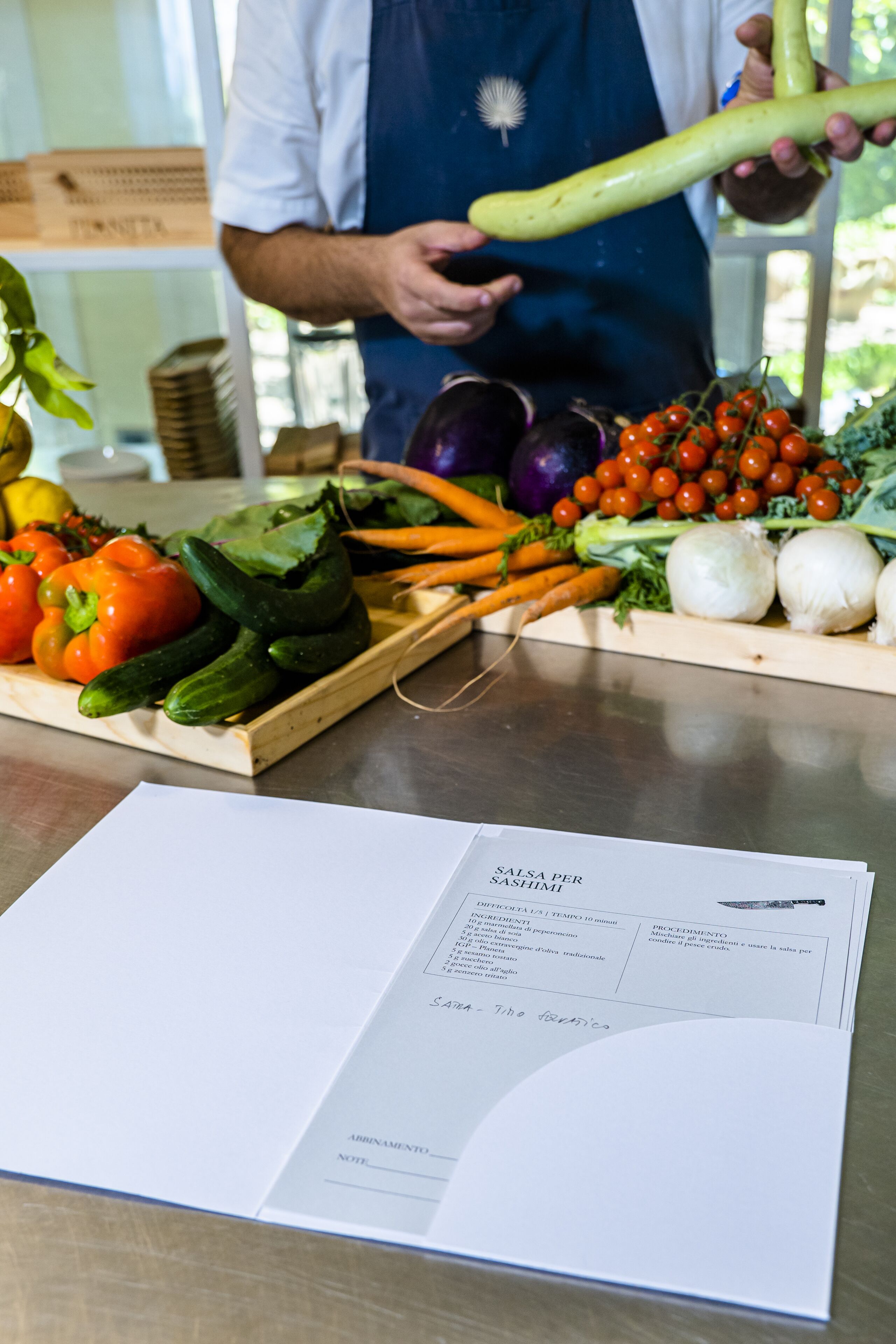 A person holding a cucumber, with various vegetables and a menu on the table.