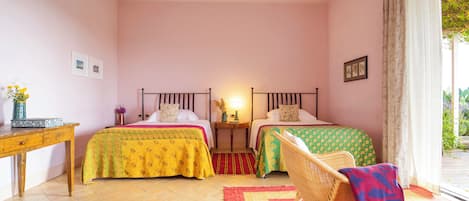 Quarto com duas camas, uma escrivaninha de madeira, uma cadeira de vime e uma janela com cortinas.
