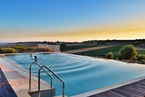 Una piscina infinita con cubierta de madera y barandillas metálicas, rodeada de colinas ajardinadas y un cielo despejado.