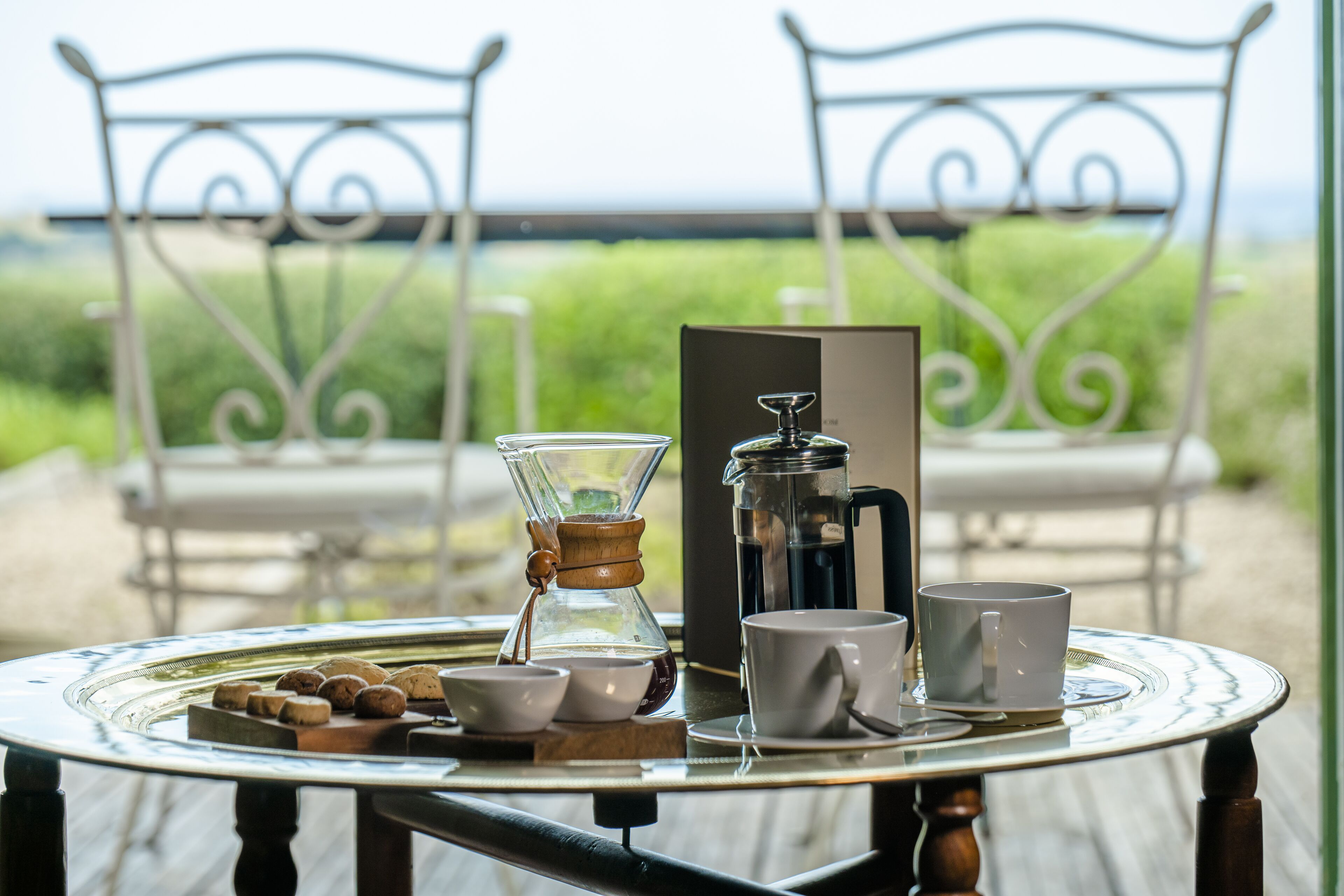 A table with a coffee maker, cups, and a tray of cookies on a deck with chairs and a view of the outdoors.