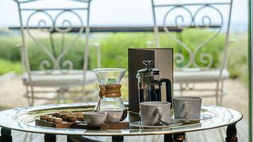 A table with a coffee maker, cups, and a tray of cookies on a deck with chairs and a view of the outdoors.